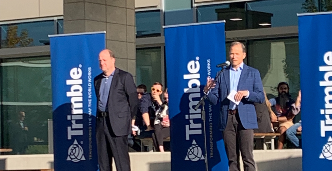 Two people presenting about the Company Trimble. The are standing in front of microphones with banners behind them.